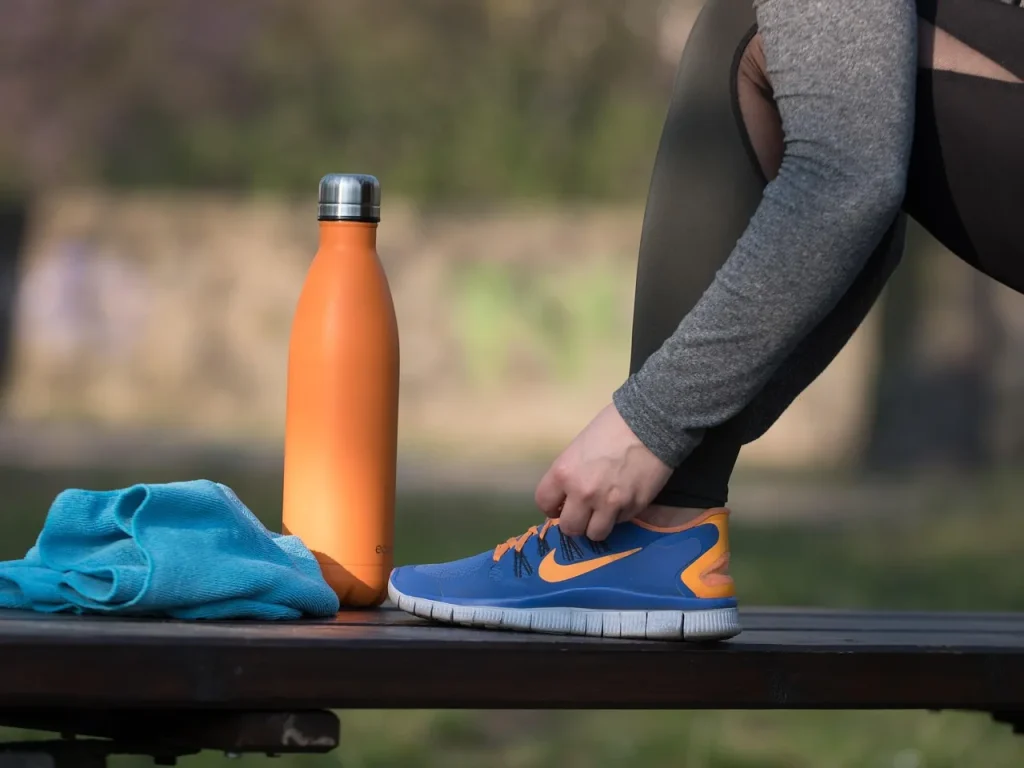 A person tying blue running shoes on a park bench next to an orange water bottle and a towel.